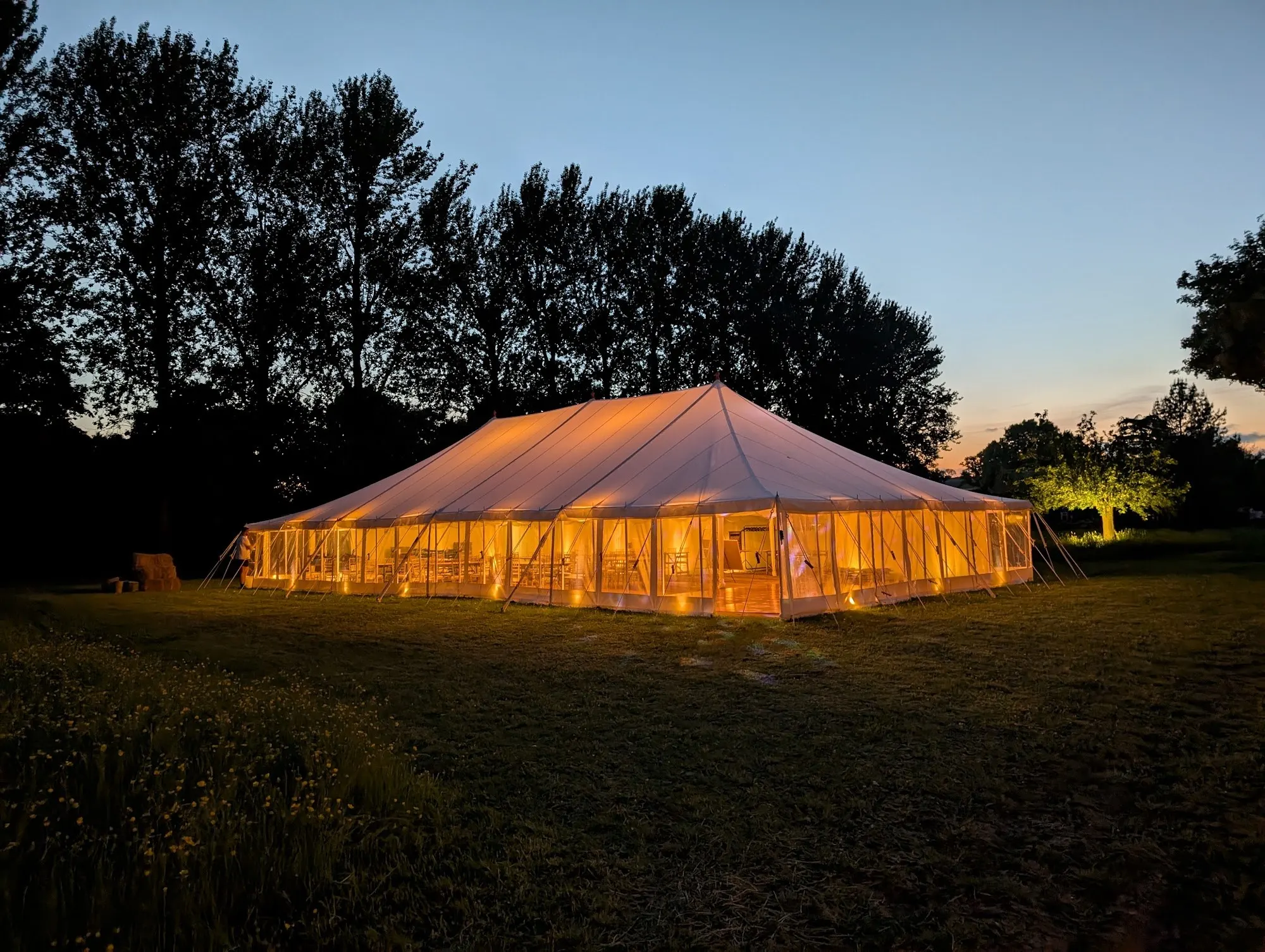 A traditional pole marquee glowing with light in a field at dusk